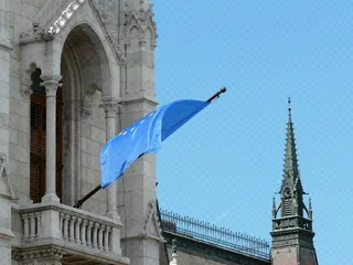 Blick auf das ungarische Parlamentsgebäude in Budapest mit einer wehenden EU‑Flagge im Vordergrund.