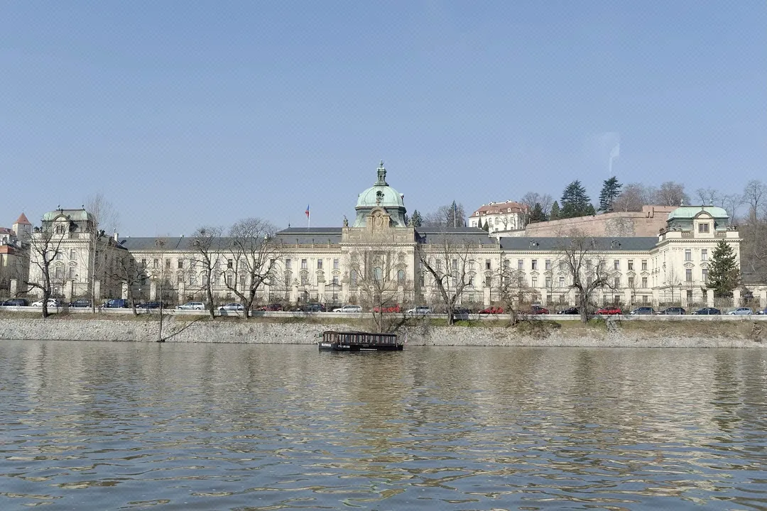 Blick über die Moldau auf die Straka-Akademie in Prag, ein großes historisches Gebäude mit hellen Fassaden und Türmen am Flussufer.
