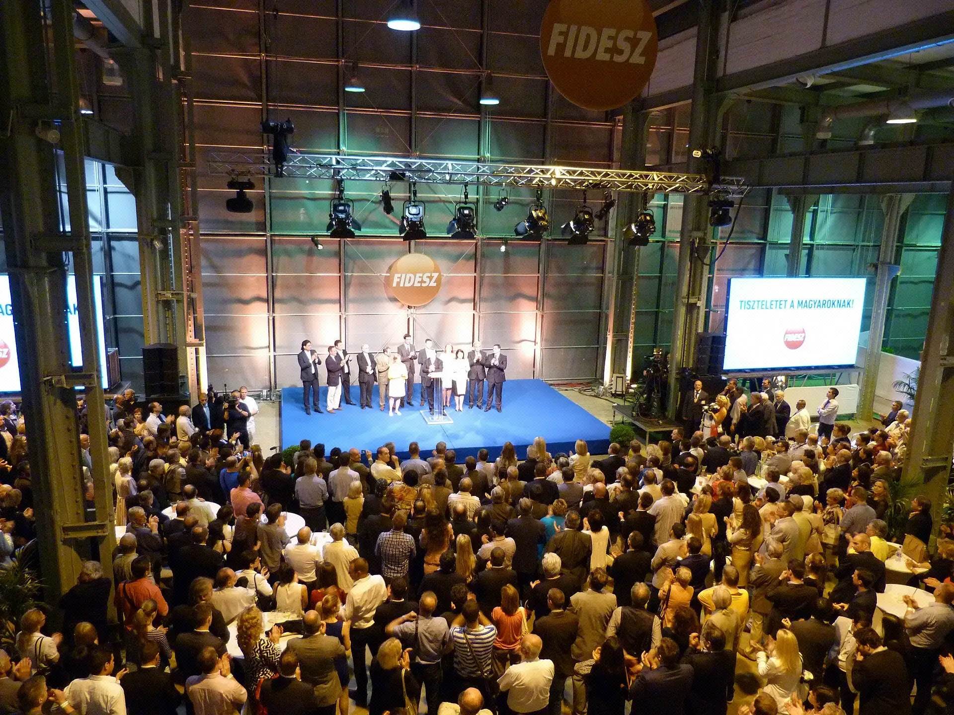 Menschen stehen dicht gedrängt in einer Halle bei einer Fidesz-Wahlveranstaltung zur Europawahl 2014 in Budapest, im Hintergrund sind Leinwände und Bühnenbeleuchtung zu sehen.